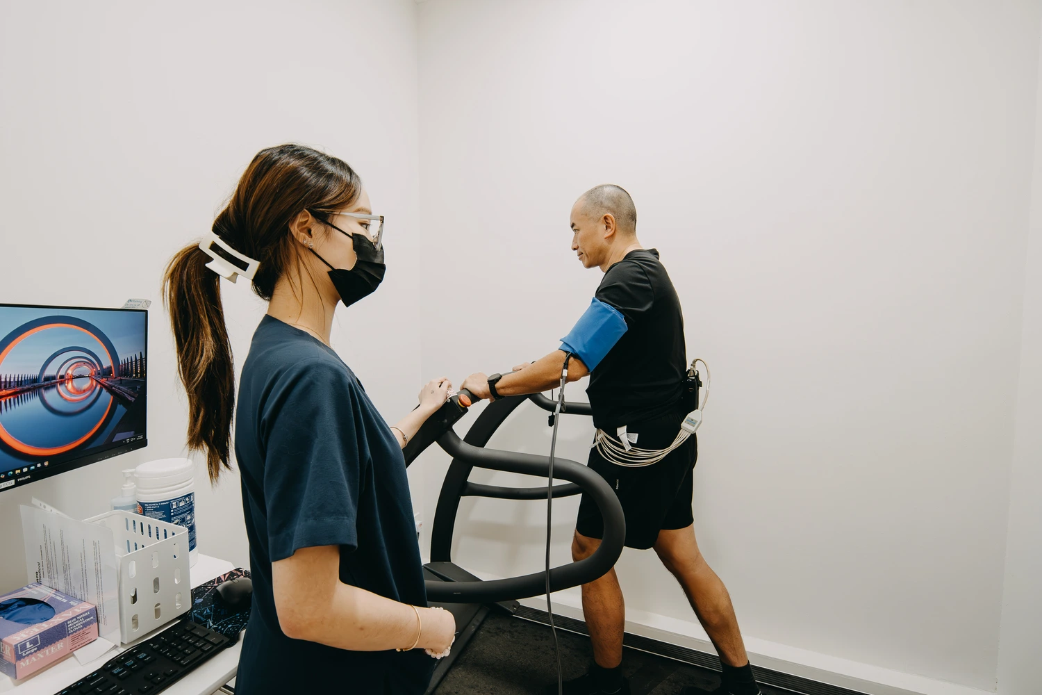 A patient undergoing a treadmill stress test in a Singapore clinic — walking on a treadmill with ECG electrodes and a blood pressure cuff while a female professional monitors the test
